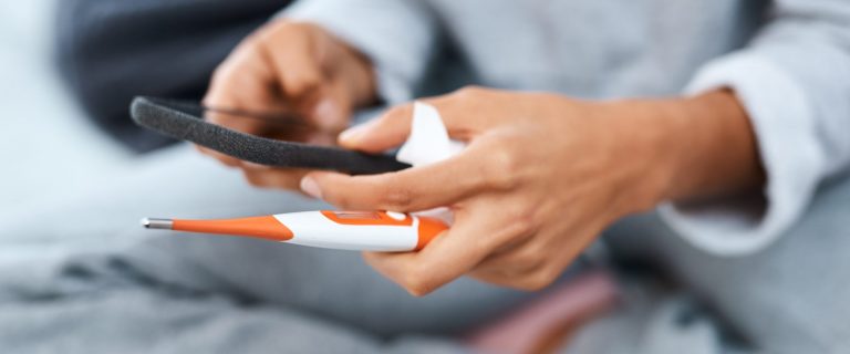 Woman using a smartphone and thermometer while recovering from an illness at home