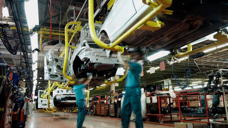 Two workers wearing green shirts and blue pants work underneath a car in an automobile factory