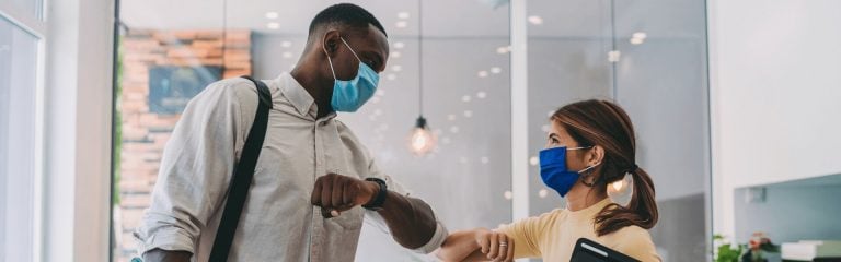 A woman and a man wearing protective masks touch each other with their elbows in greeting