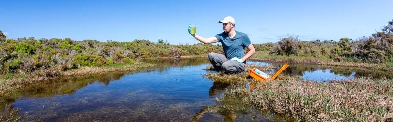 A man crouching by the water with a water sample in his hand