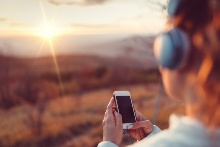 A woman is holding a phone and wearing headphones while looking out over a setting sun.
