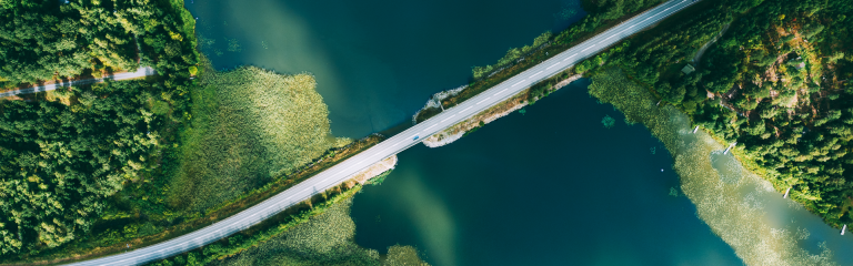 A view from above of a road spanning a body of water. Forests are visible on either side of the water and a blue car is driving across the road on the right hand side.