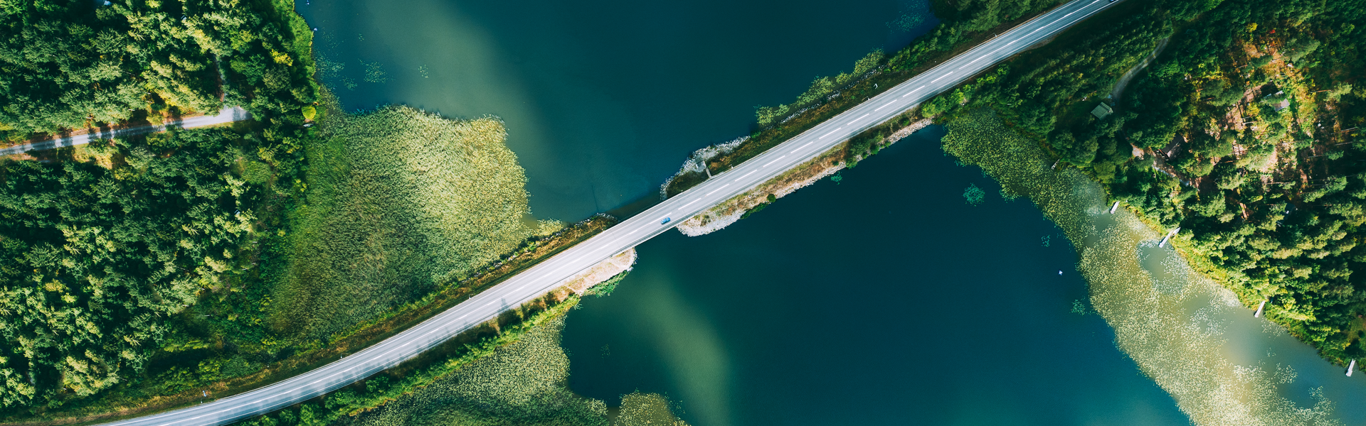 A view from above of a road spanning a body of water. Forests are visible on either side of the water and a blue car is driving across the road on the right hand side.