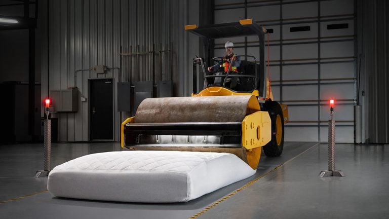 Yellow steamroller about to crush a white mattress in an industrial warehouse with red warning lights and metal walls