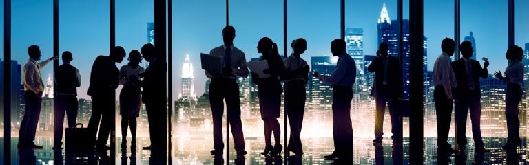 Business women and men standing against a cityscape of skyscrapers at night, discussing in groups.