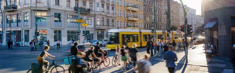 Yellow tram passing on a busy street in a European city. Pedestrians and cyclists cross at a traffic junction or wait at the lights.
