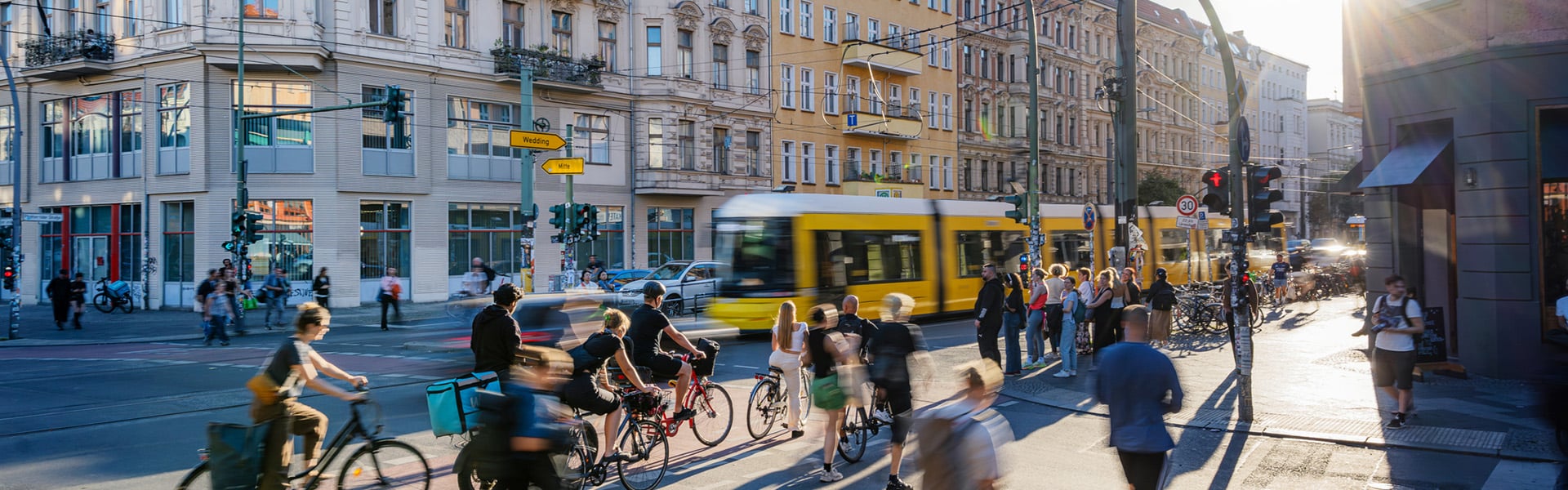 Yellow tram passing on a busy street in a European city. Pedestrians and cyclists cross at a traffic junction or wait at the lights.